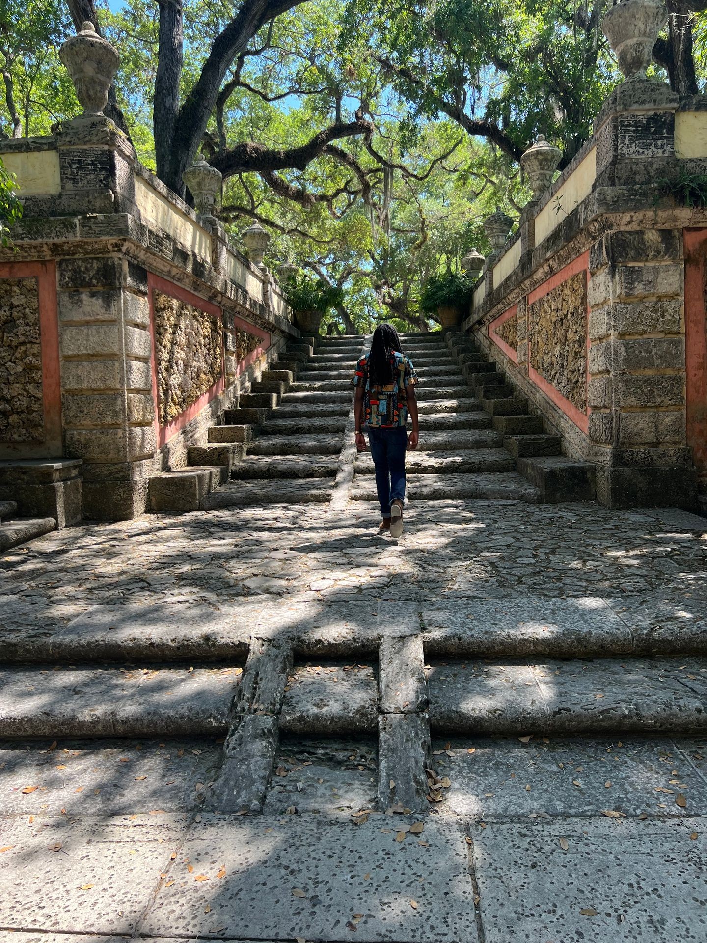 Person walking up a historical stone staircase surrounded by greenery and trees on a sunny day.