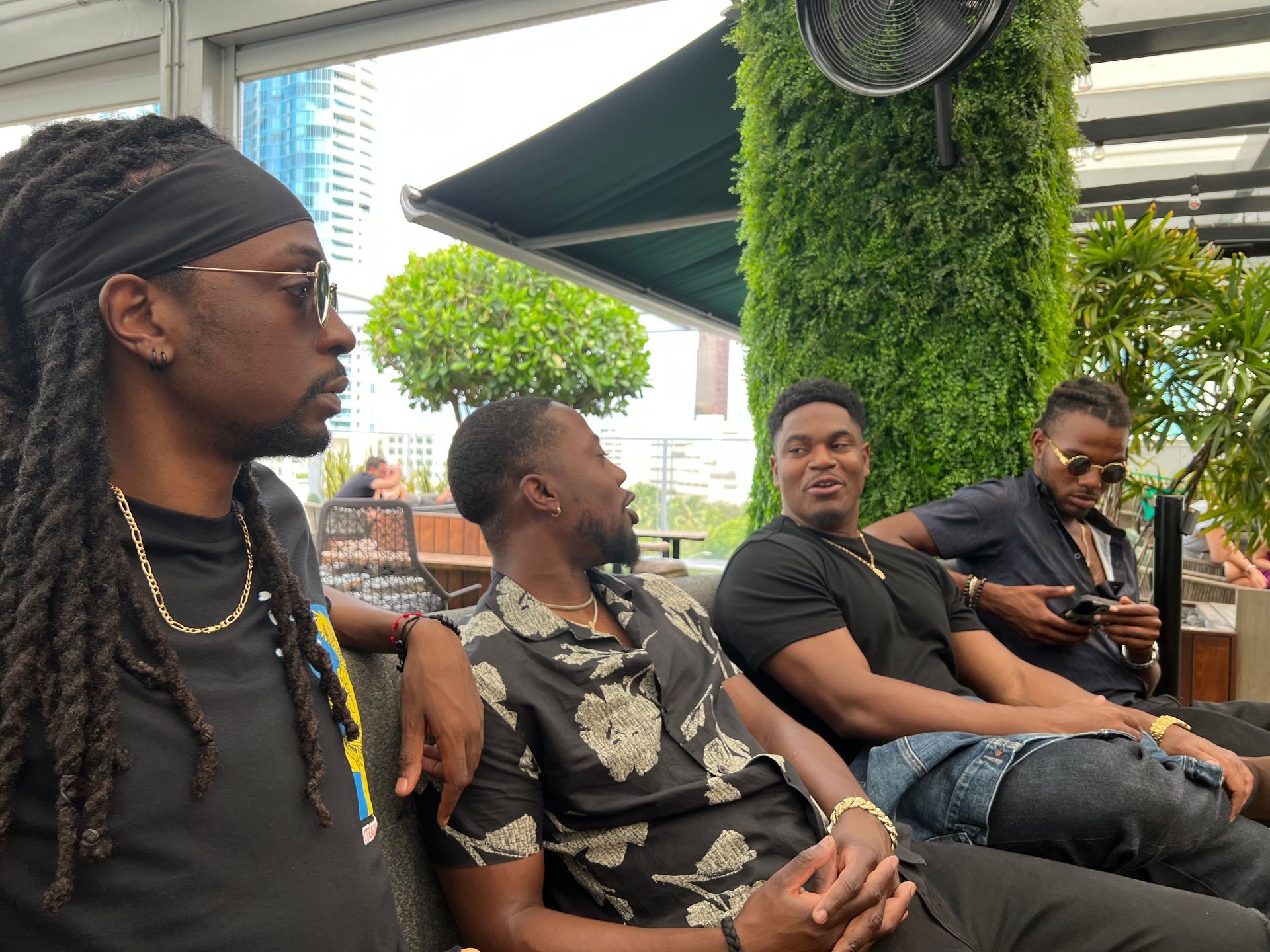 Group of men sitting outdoors in a casual setting with greenery and cityscape in the background.