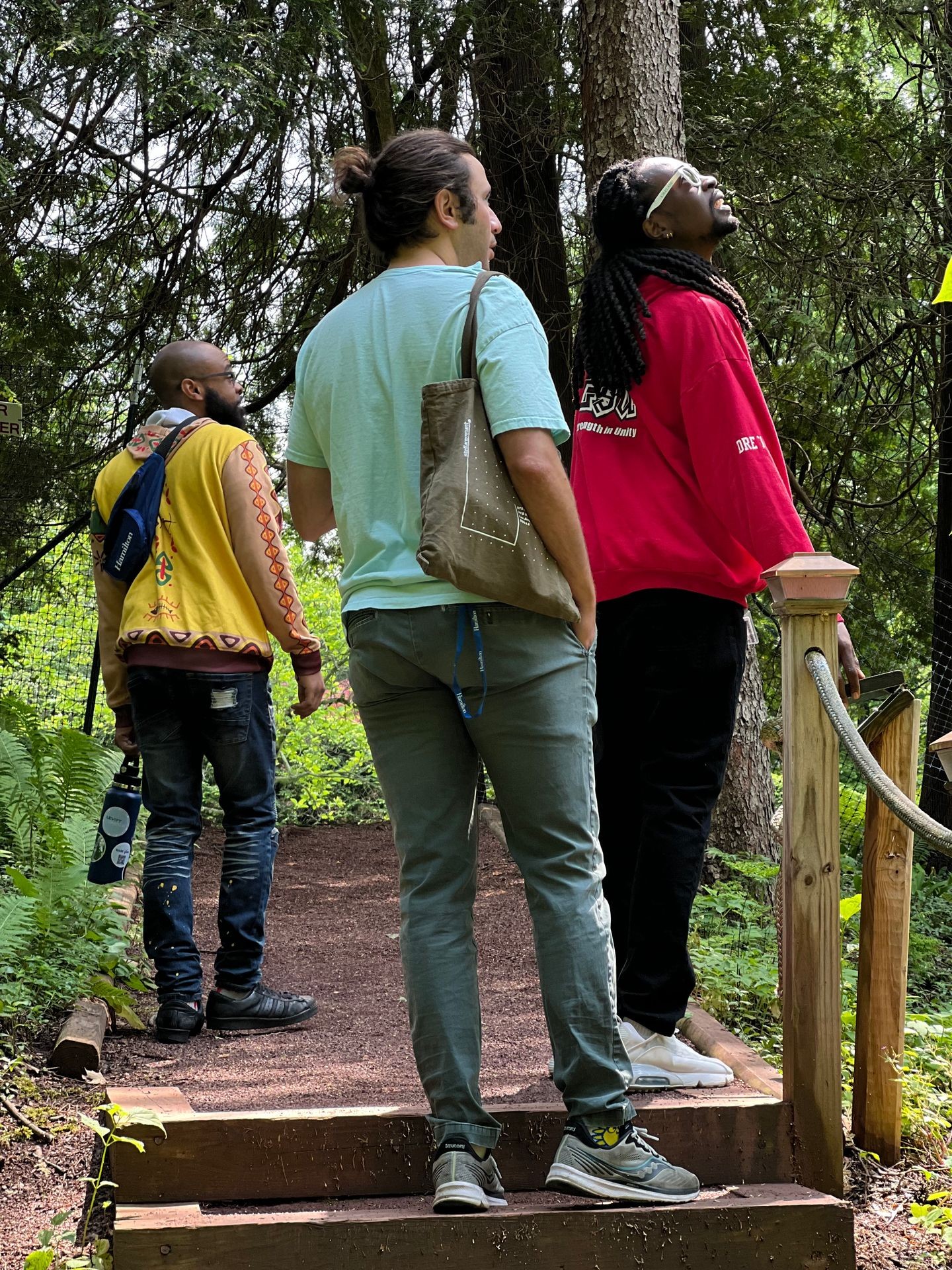 Three people standing on a forest trail, one wearing a green shirt and carrying a bag.